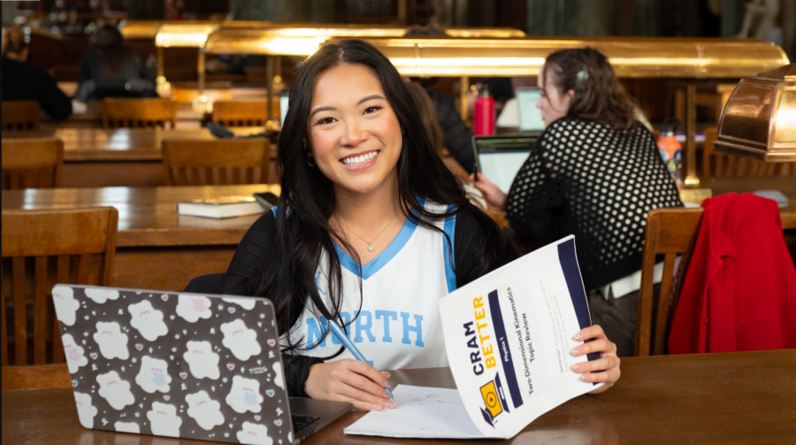 A young woman in a college classroom smiles at the camera while reading her syllabus with CramBetter on her computer